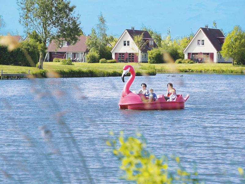 Twee personen varen met een roze flamingo waterfiets op een rustig meer met huizen op de achtergrond.