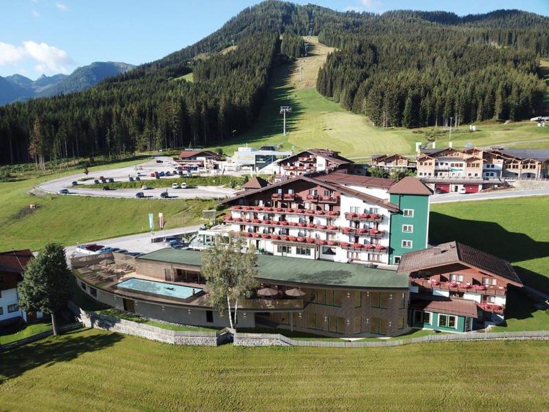 Górski ośrodek z tradycyjnymi chatami, zielonymi łąkami i zalesionymi wzgórzami w tle.