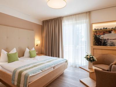 Jasny pokój hotelowy z podwójnym łóżkiem, zielonymi poduszkami, fotelem i dużym oknem z zasłonami.