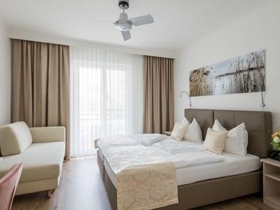 Jasny pokój hotelowy z łóżkiem podwójnym, sofą, wentylatorem sufitowym i dużym oknem z zasłonami.