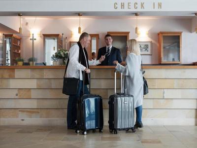 Zwei Reisende mit Koffern stehen vor dem Hotel-Check-in-Schalter, wo zwei Mitarbeiter helfen.