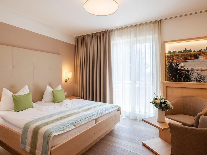 Jasny pokój hotelowy z podwójnym łóżkiem, zielonymi poduszkami, fotelem i dużym oknem z zasłonami.