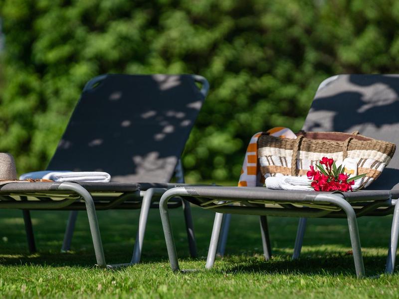 Dwa leżaki z poduszkami i kocyk na zielonej trawie, w tle krzewy.