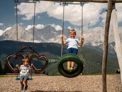 Des enfants jouent sur des balançoires dehors avec des montagnes en arrière-plan par une journée ensoleillée.