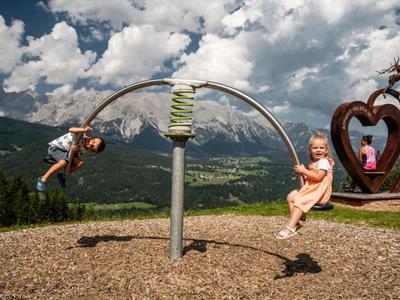 Deux enfants jouent sur un équipement de jeu moderne avec montagnes et ciel en arrière-plan.