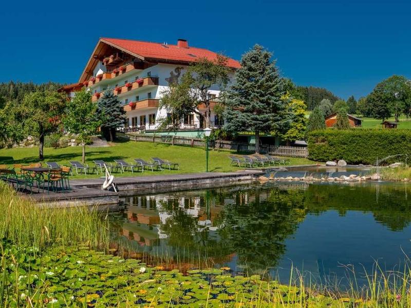 Berghotel met rood dak bij een meer, omringd door weilanden en bergen op de achtergrond.