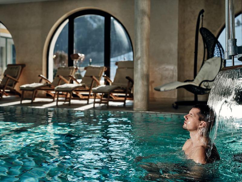 Muž relaxuje v krytém bazénu vedle vodopádu s lehátky v pozadí.