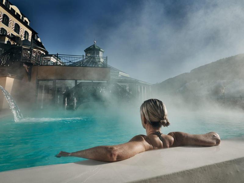 Osoba odpočívá v teplém venkovním bazénu hotelu s výhledem na hory a párou v chladném vzduchu.