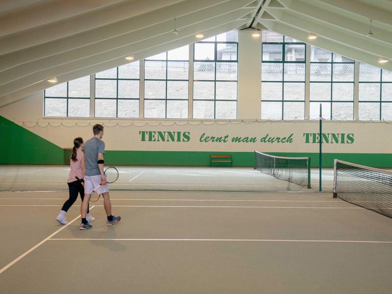 Dva lidé jdou s tenisovými raketami v kryté tenisové hale.