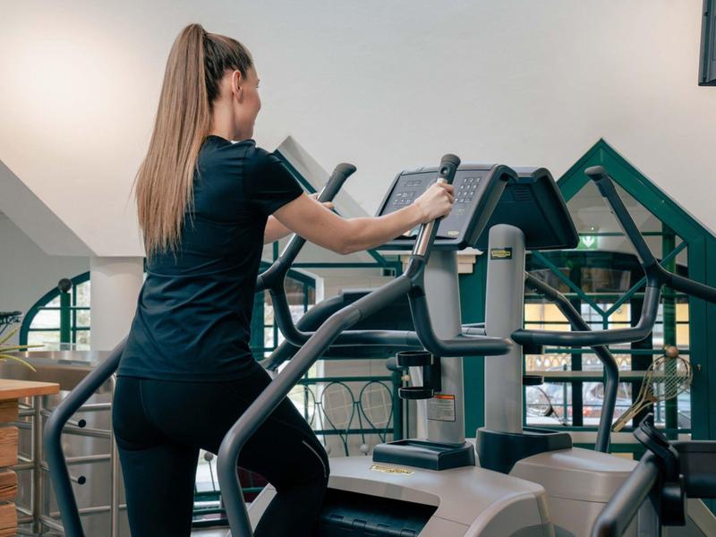 Žena cvičí na eliptickém trenažéru v dobře vybavené posilovně s velkými okny.