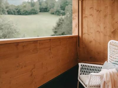 Balkon mit Holzwänden, einem weißen Korbstuhl und Blick auf eine grüne Wiese.