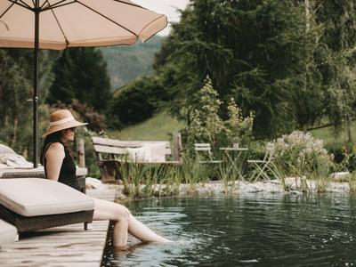 Woman sitting by a pool under an umbrella in a green, mountainous landscape.