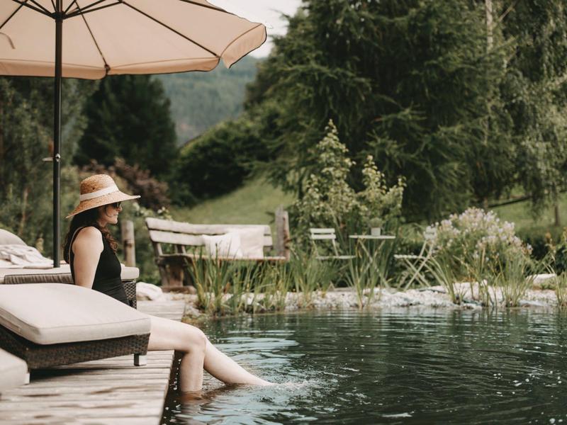 Woman sitting by a pool under an umbrella in a green, mountainous landscape.