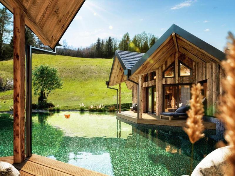 Modern wooden holiday house by water with green hill in background and clear sky.