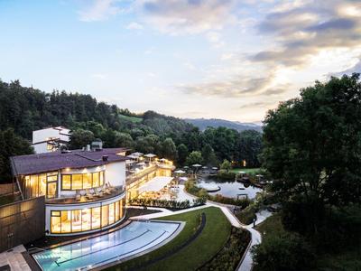 Modernes Hotel mit beleuchteten Außenpools in bewaldeter hügeliger Landschaft bei Sonnenuntergang.