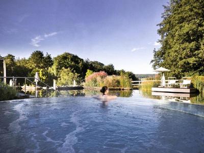 Persona relajándose en piscina exterior rodeada de árboles y cielo soleado.