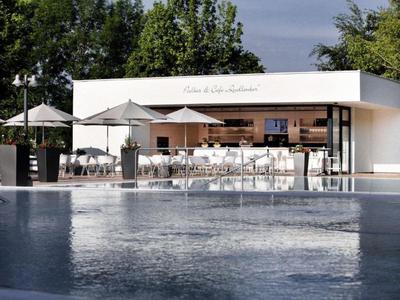 Piscina exterior moderna con bar y sombrillas blancas, rodeada de árboles verdes.