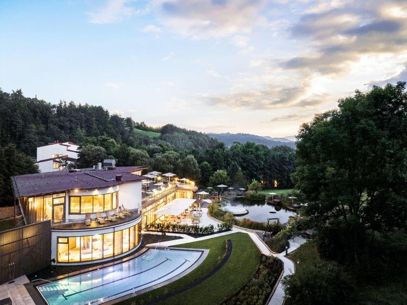 Hotel moderno con piscinas exteriores iluminadas en un paisaje boscoso y montañoso al atardecer.