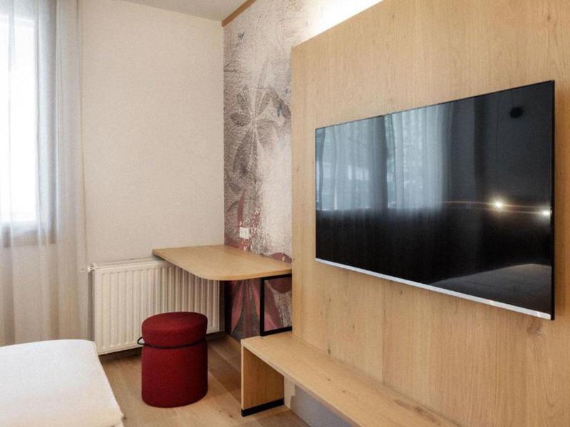 Área moderna de habitación de hotel con TV en la pared, muebles de madera y taburete rojo junto a la ventana y la cama.