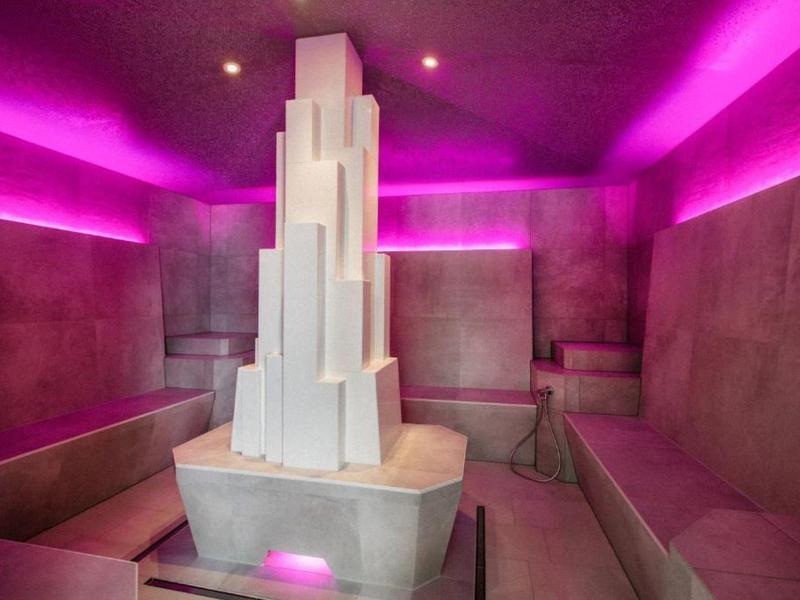 Sala de relajación moderna con bancos lounge y escultura blanca central, iluminación púrpura.