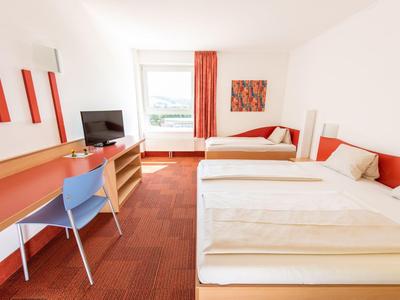 Hell eingerichtetes Hotelzimmer mit zwei Einzelbetten, Schreibtisch und Fenster mit rotem Vorhang.