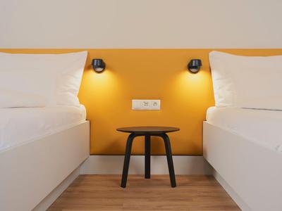 Minimalistisches Hotelzimmer mit zwei weißen Betten, gelber Wand und kleinem schwarzen Tisch.