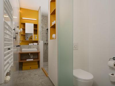Modernes Hotelbad mit Waschbecken, Toilette und gelben Regalen in kompaktem Raum.