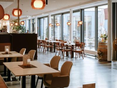 Moderner Hotel-Speisesaal mit großen Fenstern und Holzstühlen, hellem Boden und hängenden Lampen.