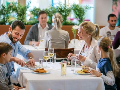 Familie genießt Mahlzeit in hellem Restaurant mit weiß gedecktem Tisch und Pflanzen im Hintergrund.