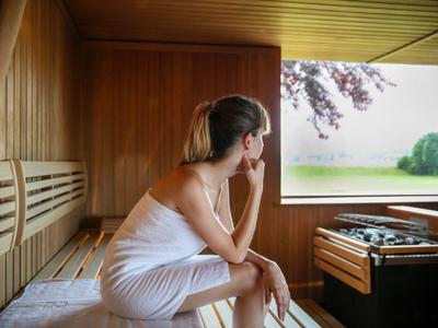 Frau in Handtuch sitzt in Holzsauna und blickt aus großem Fenster auf Natur und See.