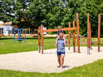 Ein Mann trägt ein Kind auf den Schultern in einem sonnigen Park mit Klettergeräten und viel Grün.