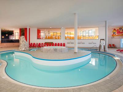 Moderner Innenpool mit geschwungener Insel, umgeben von roten Liegestühlen und großen Fenstern.