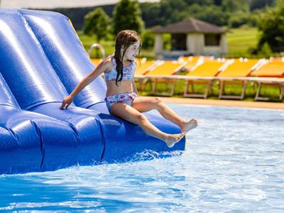 Kind spielt auf einer blauen Wasserrutsche in einem Pool mit Liegestühlen im Hintergrund.