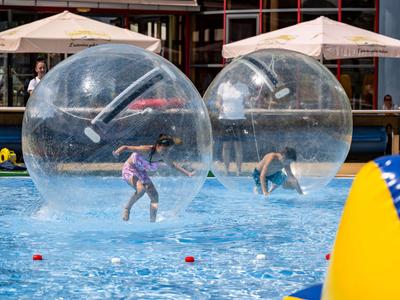 Zwei Kinder spielen in durchsichtigen Wasserbällen auf einem Pool mit Sonnenschirmen im Hintergrund.
