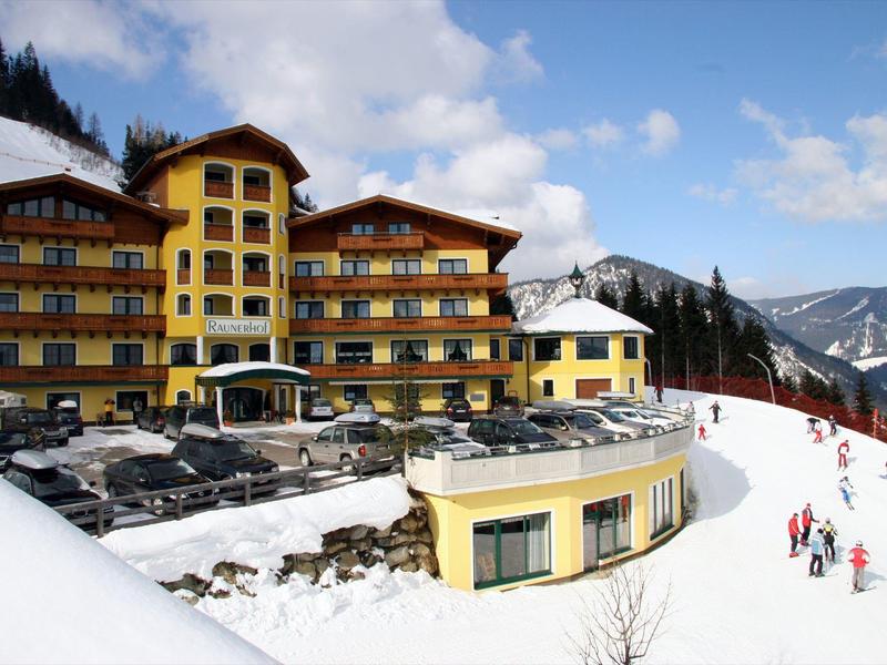 Grand hôtel avec terrasse ensoleillée dans une station de ski montagneuse enneigée sous ciel bleu.