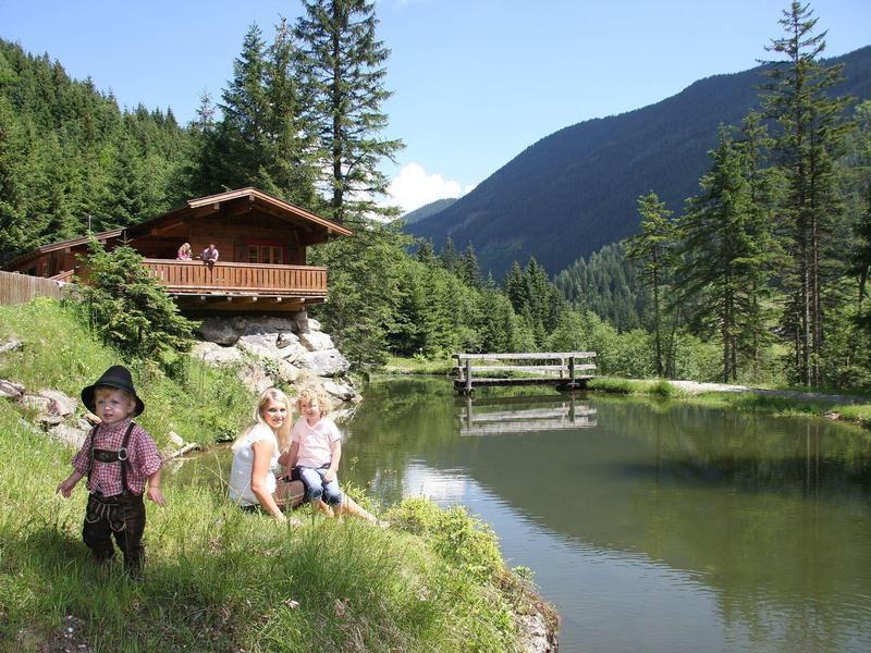 Des enfants jouent près d'un petit lac avec une cabane et des montagnes en arrière-plan.