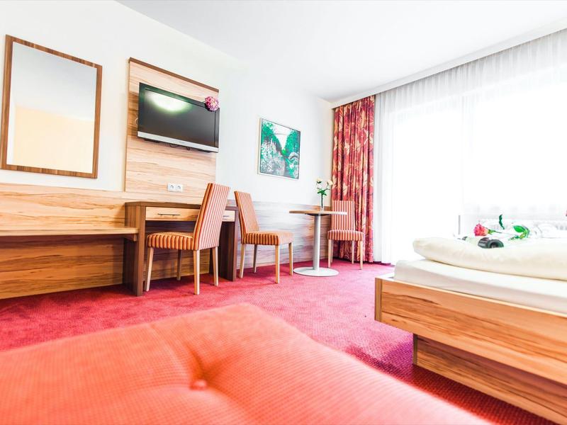 Chambre d'hôtel lumineuse avec tapis rouge, deux lits et bureau en bois près de la fenêtre avec rideaux.