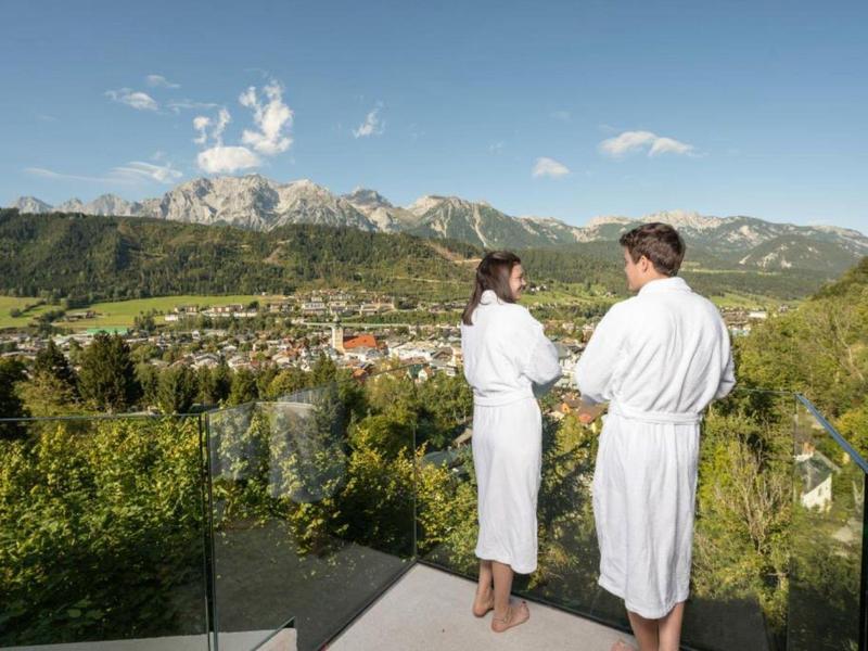 Zwei Personen in weißem Bademantel stehen auf Balkon mit Berg- und Talblick bei Sonnenschein.