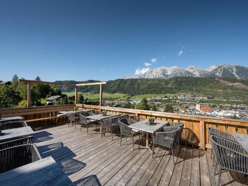Große Holzterrasse mit Stühlen und Tischen, Blick auf grüne Hügel und schneebedeckte Berge im Hintergrund.
