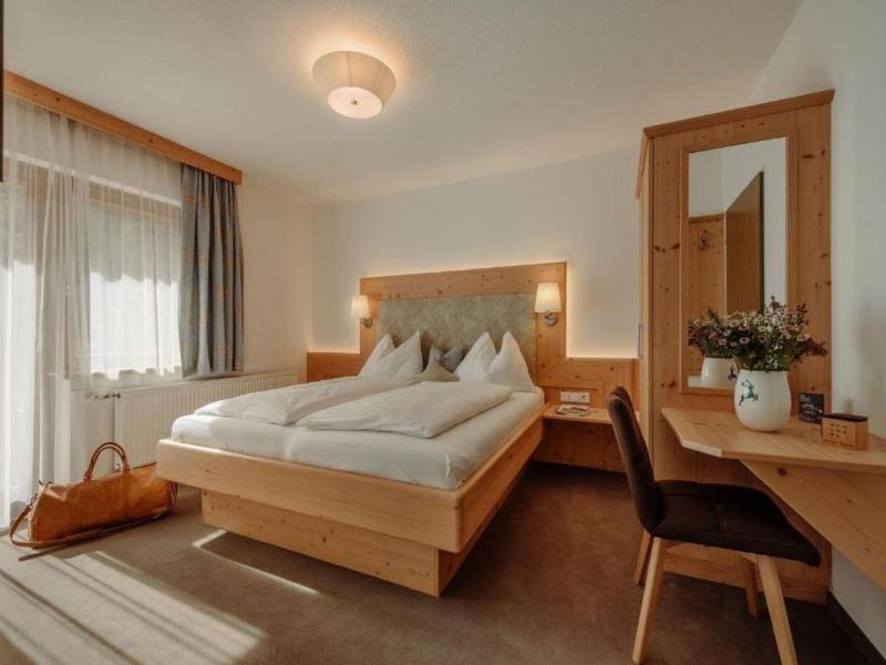 Helles Schlafzimmer mit Holzmöbeln, Doppelbett, Schreibtisch, Stuhl und Fenster mit Vorhängen.