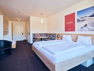 Hotelzimmer mit Doppelbett, zwei roten Postern, dunklem Teppich und Sitzbereich mit schwarzem Sessel.