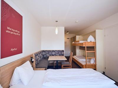 Hotelzimmer mit Etagenbett, Doppelbett, Sitzecke, Holzmöbeln und rotem Willkommensplakat an der Wand.