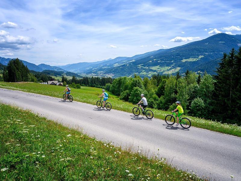 Vier Radfahrer fahren auf einer Straße durch grüne Hügel mit Bergen und blauem Himmel im Hintergrund.