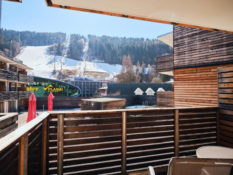 Balkon mit Holzbalken, Tisch mit Stühlen, Blick auf schneebedeckte Skipisten und umliegende Holzgebäude.