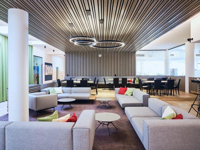 Modernes, helles Büro mit grauen Sofas, bunten Kissen, runden Tischen und Holzdecken mit Lichtringen.