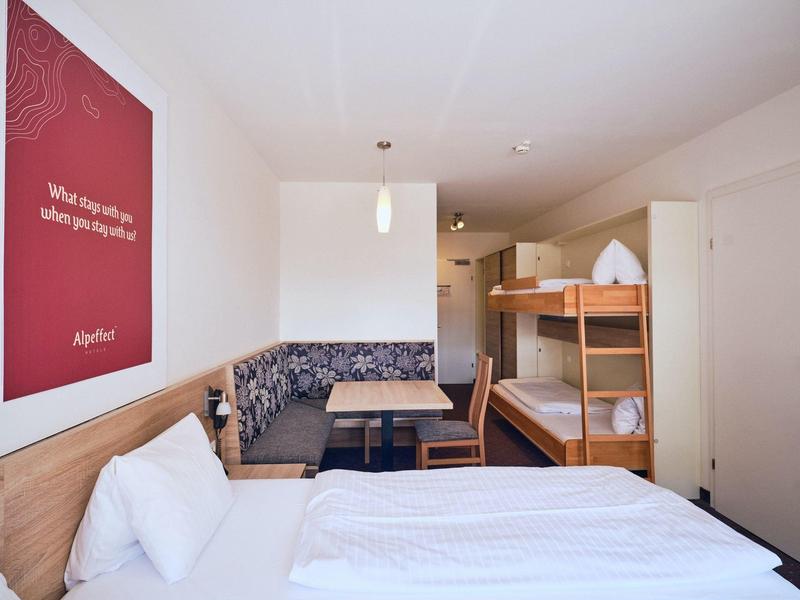 Hotelzimmer mit Etagenbett, Doppelbett, Sitzecke, Holzmöbeln und rotem Willkommensplakat an der Wand.