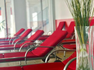 Rote Liegestühle in einem hellen Innenbereich, im Vordergrund eine Vase mit grünen Gräsern.