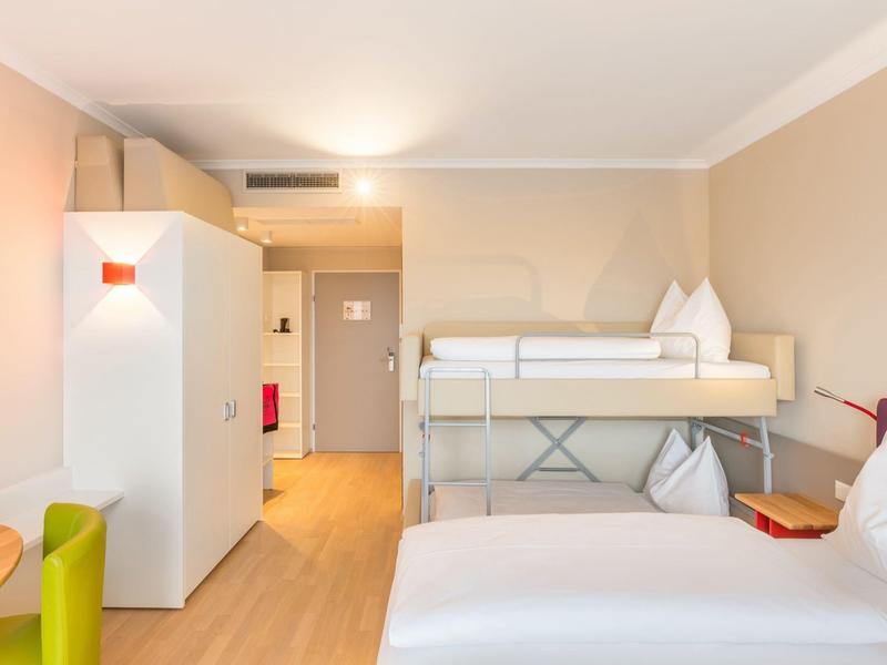 Helles Hotelzimmer mit Doppelbett, Etagenbett, Holzfußboden und Blick zum Flur.