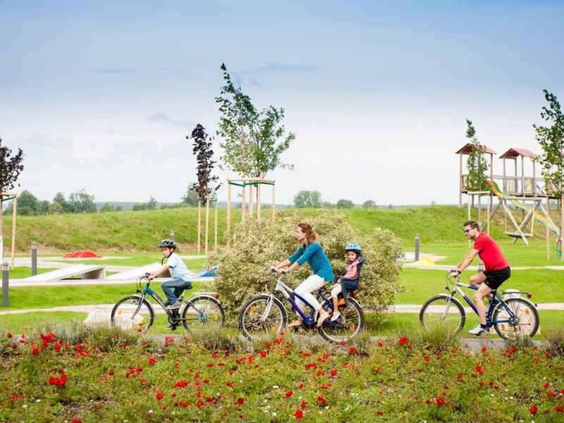 Familie fährt mit Fahrrädern auf einem landschaftlichen, blumenreichen Radweg neben einem Spielplatz.