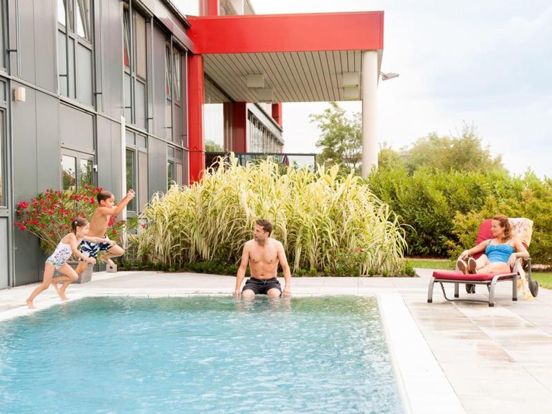 Ein Kind springt in einen Pool, während zwei Erwachsene am Poolrand entspannen.